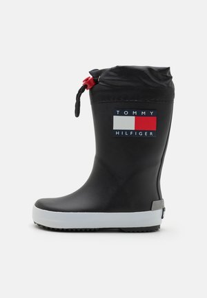 Bota de lluvia de goma negra con suela gris-blanca, con una parte superior de tela negra y un cordón rojo. Muestra un logotipo multicolor en un lado.