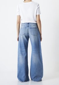 Persona con jeans claros de pierna ancha y una camiseta blanca de manga corta, de pie con la espalda hacia la cámara.