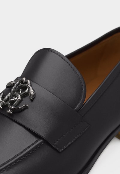 Čierna kožená mokasína s hladkým povrchom, viditeľným prešívaním a strieborným dekoratívnym kovovým emblémom na remienku, s viditeľou koňakovou vnútornou podšívkou.