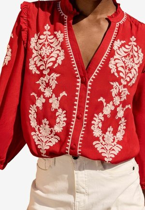 Red long-sleeve blouse with white floral embroidery, buttoned front, and ruffled collar, worn with high-waisted white pants.
