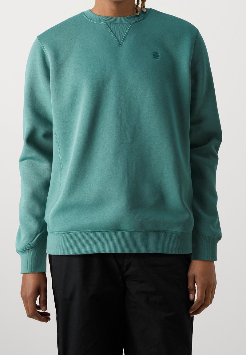 Personne portant un sweat-shirt col rond turquoise avec un petit logo sur la poitrine et un pantalon noir, debout devant un fond gris uni.