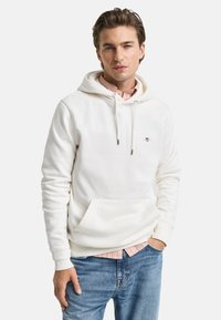 White hooded sweatshirt with a front pocket, drawstrings, and small embroidered logo. Worn over a light plaid shirt, paired with blue jeans.