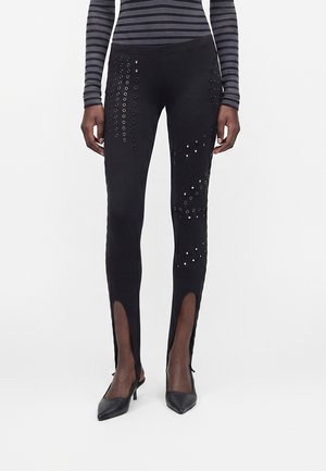 Czarne spodnie z dekoracyjnymi oczkami w wzory na udach i łydkach, z przednimi rozcięciami nad spiczastymi czarnymi obcasami, noszone z paskowanym topem.