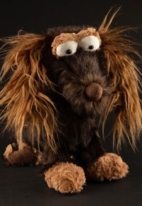 Stuffed toy dog with a brown and black plush body, long wavy fur, large white eyes, and a round brown nose; fuzzy paws complete the design.