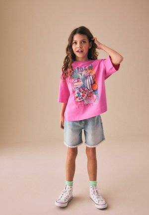 Pikkade laineliste juustega tüdruk, kes kannab erkroosat "Garden Party" särki, teksapükse, rohelisi sokke ja valgeid kõrge säärega tossusid, seistes käsi pea lähedal.