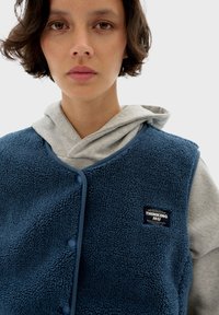 Niebieska kamizelka polarowa z zapięciem na guzik i małą metką marki. Noszona na szarym hoodie z troczkiem i miękką fakturą.