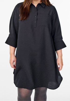 Femme portant une robe chemise noire ample avec des manches trois-quarts, un col boutonné, une poche poitrine et des collants gris foncé.