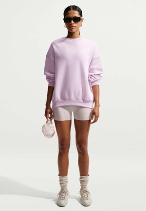Donna che indossa una felpa oversize viola chiaro, pantaloncini da ciclista beige, calzini arricciati, sneakers, occhiali da sole, tenendo una piccola borsa rotonda.