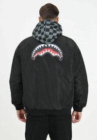 Giacca bomber nera con una grande grafica di una bocca di squalo sul retro, dotata di un cappuccio grigio a scacchi e polsini e fondo a coste.