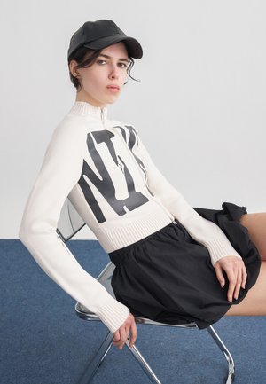 Joven con gorra negra, suéter blanco con cremallera y letras negras, y falda negra, sentada en una silla moderna de cromo sobre una alfombra azul.