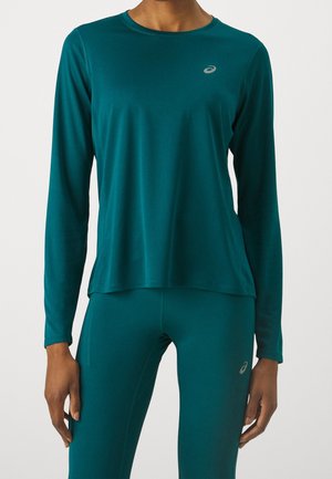 Personne portant un haut de sport à manches longues bleu sarcelle et des leggings assortis, tous deux avec de petits logos Asics gris clair sur la poitrine et la cuisse.