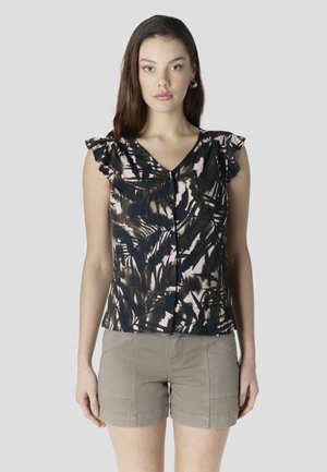 Joven de pie, vestida con una blusa oscura con estampado de hojas y mangas con volantes, y pantalones cortos beige, mirando al frente contra un fondo liso.