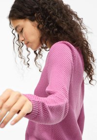 Sweter dziergany na drutach w jasnoróżowym kolorze z fakturowanym wzorem, okrągłym dekoltem i długimi rękawami. Posiada ściągacze na mankietach oraz luźny krój.