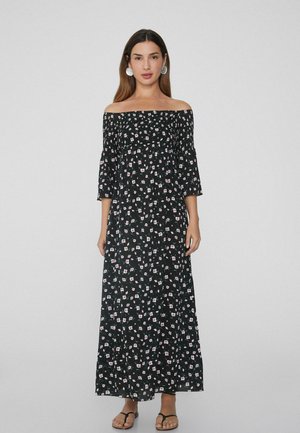 Vestido maxi de tela negra con estampado floral en blanco y rosa, diseño de hombros al descubierto, mangas 3/4 y un ajuste suelto y fluido. Sandalias planas.