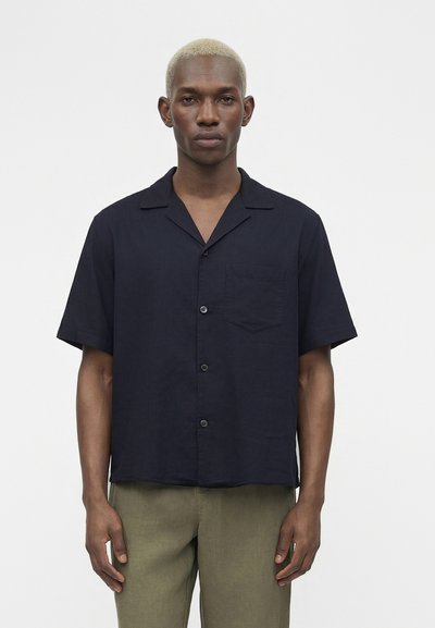 Homme aux cheveux blonds courts, portant une chemise noire à manches courtes boutonnée et un pantalon vert olive, debout devant un fond uni.