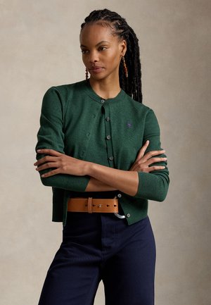 Femme aux longs cheveux tressés porte un cardigan vert boutonné, une ceinture marron, un pantalon bleu marine et des boucles d'oreilles en ambre, debout les bras croisés.