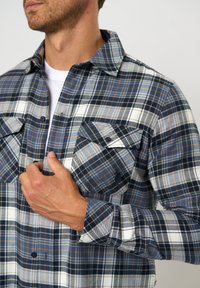 Mees, kes kannab sinise, valge ja oranži ruudulise mustriga nööbitavat särki valge t-särgi peal, hoides särki vasaku käega rinna juures.