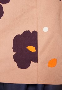 Tecido com padrão floral em tom areia, com detalhes em púrpura escuro e laranja. Textura suave, com uma costura proeminente ao longo da borda.