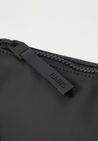 Schwarze wasserdichte Tasche mit einem strukturierten Reißverschluss und einem flachen, stabilen Reißverschlussgriff, der mit "RAINS" beschriftet ist. Glatte, matte Oberfläche mit minimalistischer Gestaltung.