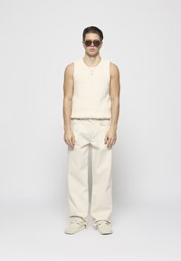 Strickwaren in Creme ohne Ärmel mit Zopfmuster, kombiniert mit hellbeigen weiten Hosen und passenden weichen Wildlederschuhen.