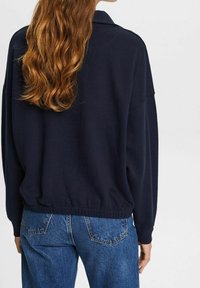 Sudadera de gran tamaño en azul marino con dobladillo y cuello acanalados, combinada con pantalones vaqueros de tiro medio que cuentan con bolsillos traseros y una textura desgastada.