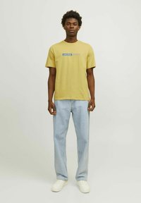 Camiseta de algodón amarilla con un texto de logo azul, combinada con jeans de mezclilla azul claro y zapatillas blancas, de pie contra un fondo neutro.