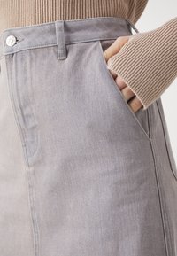 Fustă din denim gri cu o textură netedă, buzunare laterale și un sistem de închidere vizibil cu un buton metalic la talie. Prezintă o croială clasică, adaptată.