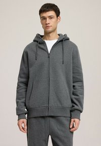 Szara bluza z kapturem na zamek, wykonana z miękkiego materiału, z kapturem ze sznurkiem, przednimi kieszeniami oraz ściągaczami przy mankietach i u dołu. Minimalistyczny design z detalem logo.