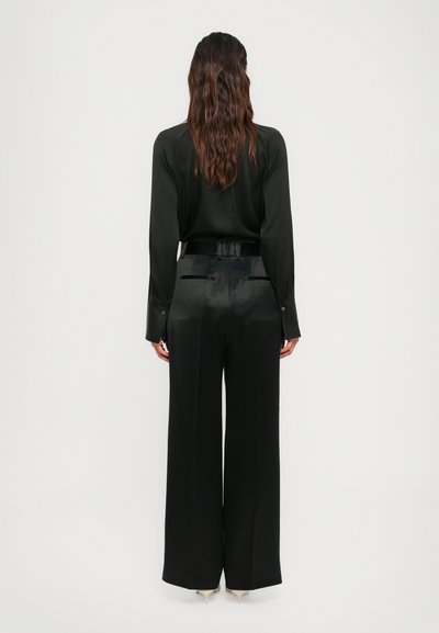 Woman with long wavy hair wearing a black long-sleeve shirt and high-waisted wide-leg black pants with beige heels, standing facing away.