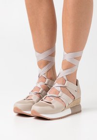 Baskets compensées beiges avec des sangles croisées en ruban, en matériaux en maille et cuir, surface texturée et semelle en caoutchouc avec des accents blancs et marron.
