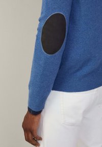 Sininen neulepusero, jossa on musta nahkalaikkupatchi kyynärpäissä, ribatulla hihalla ja sileällä pinnalla. Valkoiset housut näkyvät osittain.