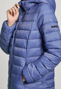 Vadderad jacka i ljusblått med dragkedja. Har horisontella sömmar och en logotypapplikation på ärmen. Mjuk, slät textur.