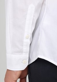 Puño de camisa de lino blanco con dos botones, con una textura suave y un corte preciso. La tela muestra un tejido sutil y ligero.