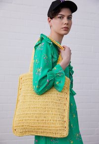 Persoon in groen bloemenjurk en zwarte pet houdt grote gele geweven schoudertas over schouder tegen een witte bakstenen muur.