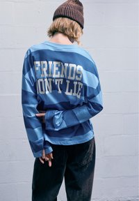 Dlhý rukáv s košeľou, ktorá má modré horizontálne pruhy, na chrbte s textom "PRIATELIA NEKLAMÚ" v bielej farbe. V kombinácii s tmavými nohavicami.