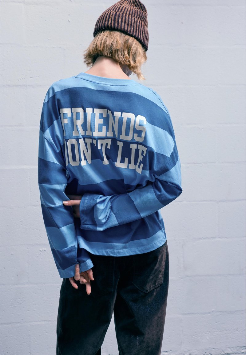 Dlhý rukáv s košeľou, ktorá má modré horizontálne pruhy, na chrbte s textom "PRIATELIA NEKLAMÚ" v bielej farbe. V kombinácii s tmavými nohavicami.