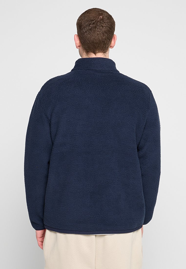Pullover in pile blu navy con colletto alto, tessuto texturizzato e vestibilità rilassata, caratterizzato da un colore uniforme e senza loghi o motivi visibili.