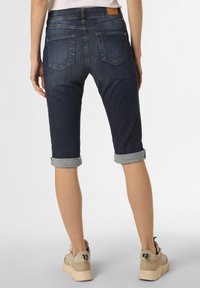 Dunkelblaue Capri-Jeans mit aufgerollten Säumen, Gesäßtaschen und einem Lederpatch. Getragen mit beigen Sneakern vor einem hellen Hintergrund.