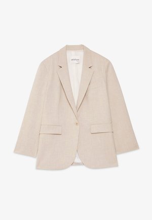 Ung kvinde med langt mørkt hår iført beige jakkesæt med vide bukser og enkeltknappet blazer over hvid top, stående på neutral baggrund.