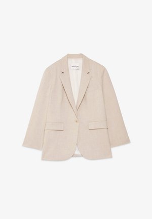 Giovane donna con lunghi capelli scuri che indossa un completo beige con pantaloni a gamba larga e blazer monopetto sopra una maglia bianca, in piedi su uno sfondo neutro.