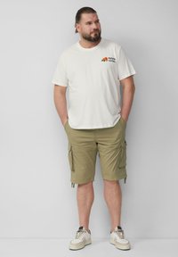 Man met een baard en samengebonden haar, gekleed in een wit T-shirt met opdruk, kaki cargoshorts en witte sneakers, staand met zijn handen in zijn zakken.