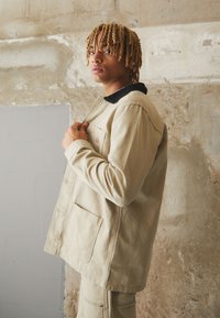 Beige Canvas-Arbeitsjacke mit einem schwarzen Cordkragen, seitlichen Taschen und einem kleinen Logopatch. Modell posiert an einer strukturierten Betonwand.