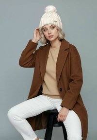 Brown wool coat over a beige sweater, white pants, and a cream knit beanie with a pom-pom, sitting on a black stool against a gray backdrop.