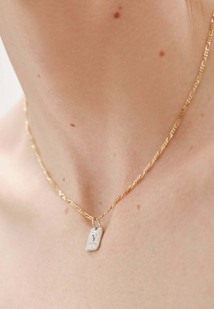 Collar de cadena de oro con un pequeño colgante de plata grabado con la letra "V", llevado en el cuello de una persona con lunares visibles en la piel.