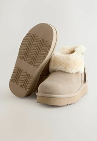 Beige Wildleder-Stiefeletten mit einem flauschigen, cremefarbenen Kragen, flacher Plateau-Sohle und strukturiertem Gummi-Außensohle mit Zickzackmuster.