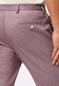 Helllilafarbene Hose aus strukturiertem Material, mit einer Seitentasche mit Knopfverschluss und einem kleinen Markenetikett an der Gesäßtasche.