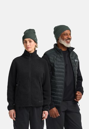Een vrouw en een man dragen donkergroene beanies en zwarte outdoorjacks, staan zij aan zij tegen een witte achtergrond.