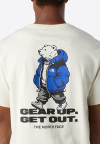 Postava ledního medvěda oblečená v modré péřové bundě a teniskách, která jde s nápisem "PŘIPRAV SE. VYJDI VEN. THE NORTH FACE" na bílé košili zezadu.