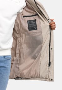 Beige outer jacket with a smooth texture, featuring a zipper and snap closures. Inside, a light satin lining and a label detailing care instructions.