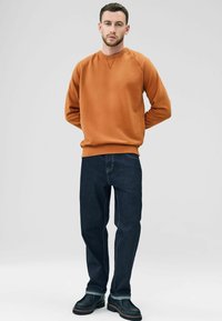 Sudadera naranja con cuello redondo y puños de canalé, combinada con jeans de mezclilla azul oscuro con costuras en contraste y botas negras.